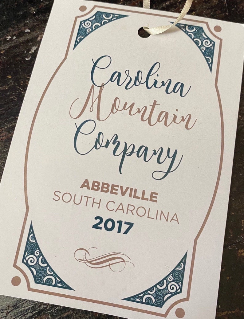 May include: A white tag with brown and blue decorative borders. The tag has the text "Carolina Mountain Company" in cursive script, followed by "Abbeville, South Carolina 2017".