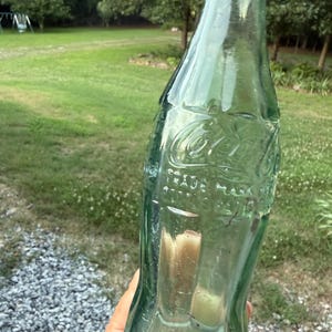 Puede incluir: Una botella de vidrio verde claro vintage de Coca-Cola con forma contorneada. El vidrio presenta letras en relieve que dicen Coca-Cola, Trade Mark Registered, y la botella tiene un cuello estrecho y una base ancha con residuos visibles.