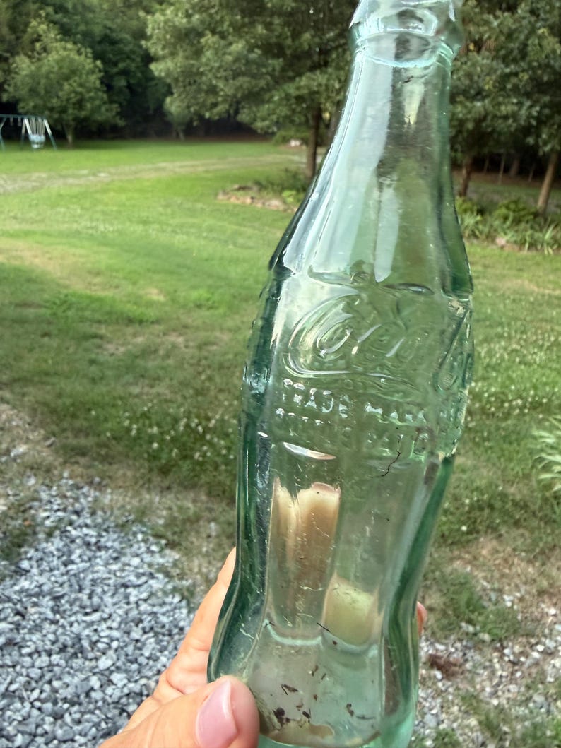 Puede incluir: Una botella de vidrio verde claro vintage de Coca-Cola con forma contorneada. El vidrio presenta letras en relieve que dicen Coca-Cola, Trade Mark Registered, y la botella tiene un cuello estrecho y una base ancha con residuos visibles.