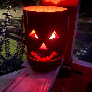 May include: A red metal jack-o'-lantern with a lit candle inside. The jack-o'-lantern has a traditional carved face with a wide smile, triangular eyes, and a triangular nose.