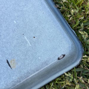 Puede incluir: Primer plano de una bandeja rectangular de esmalte azul claro con un borde azul oscuro. La bandeja tiene una superficie moteada con pequeñas manchas oscuras y algunos arañazos. La bandeja está sobre un fondo de hierba verde.