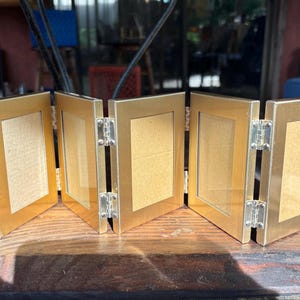 May include: A gold-colored, multi-panel picture frame with silver hinges. The frame is made of a metallic material and is divided into five sections. Each section has a rectangular opening for displaying photos or artwork.
