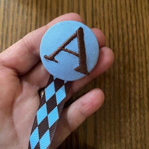 May include: A blue fabric button with a brown embroidered letter A. A brown and blue ribbon with a diamond pattern is attached to the button.