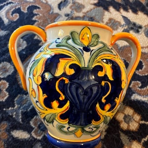 Puede incluir: Jarrón de cerámica con dos asas naranjas y un diseño floral detallado. El jarrón presenta un borde y una base blancos con detalles azules, amarillos y verdes. El cuerpo del jarrón es principalmente azul oscuro.