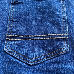 Op de afbeelding: Een close-up van een blauwe denim achterzak met een bruin stikseldetail en twee zilveren kettingen aan de bovenkant.