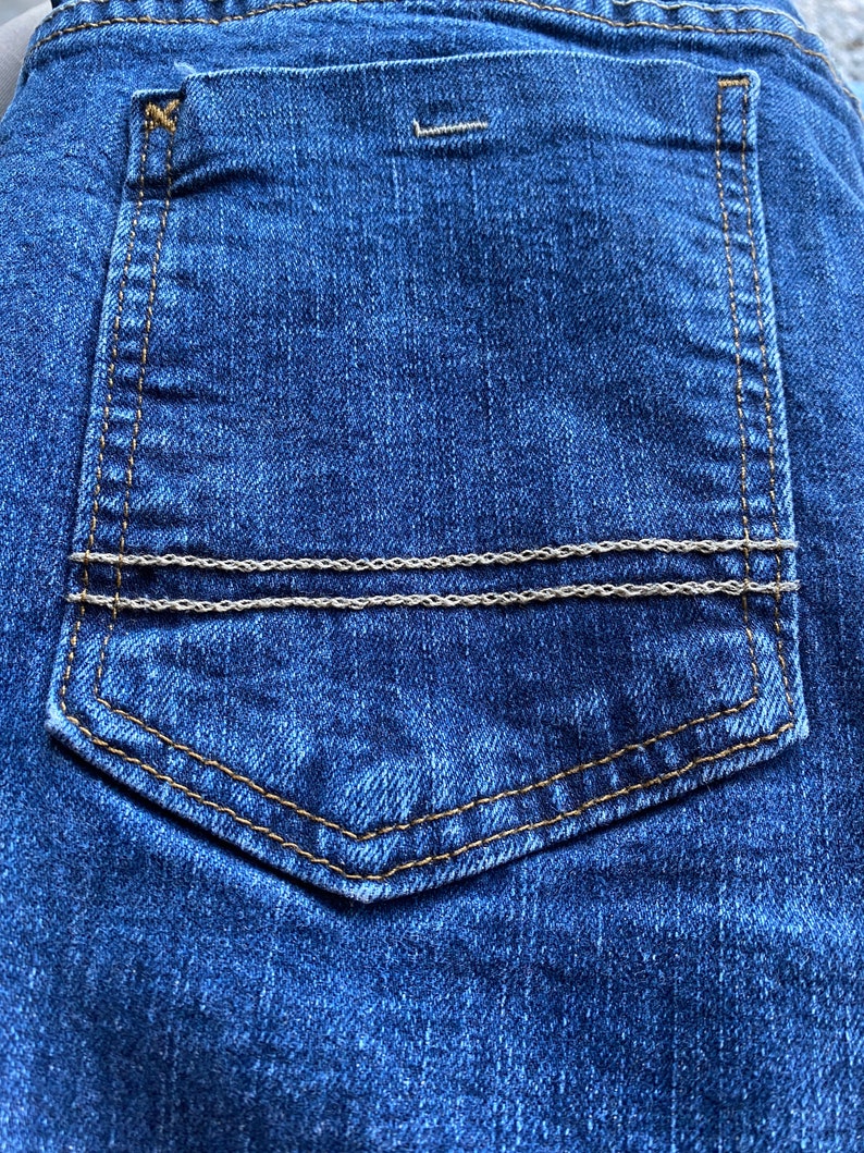 Op de afbeelding: Een close-up van een blauwe denim achterzak met een bruin stikseldetail en twee zilveren kettingen aan de bovenkant.