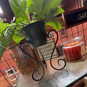 May include: A decorative metal plant stand with a potted plant, a candle in a glass jar, and a sign that reads "Blessed Beyond Measure." The stand has a scrolled design and is on a glass table. A sign that reads "Garden" is in the background.