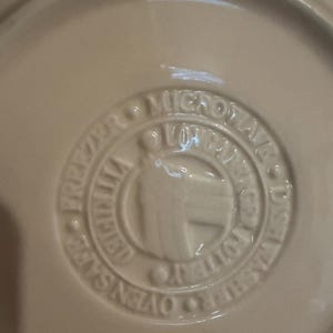 May include: A cream-colored ceramic pottery base featuring an embossed circular stamp. The text reads MICROWAVE, DISHWASHER, OVEN SAFE, FREEZER, and LONGABERGER POTTERY. The center of the stamp displays a stylized logo with the word ORIGINAL.