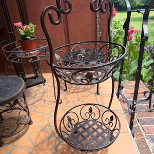 May include: A dark brown metal two-tier plant stand featuring a decorative arched handle at the top. Each tier consists of a circular basket with a lattice-patterned base and small metal leaf accents along the curved rim, supported by thin legs.