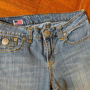 May include: Close-up of light blue denim jeans with yellow stitching. Features include a small American flag patch, metal rivets, and a button closure. The jeans have front pockets and a coin pocket.