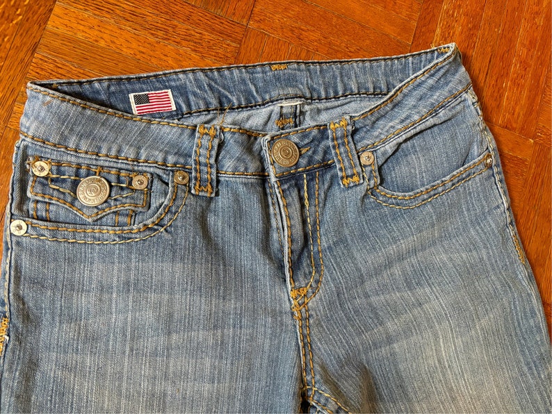 May include: Close-up of light blue denim jeans with yellow stitching. Features include a small American flag patch, metal rivets, and a button closure. The jeans have front pockets and a coin pocket.