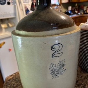 May include: A vintage brown and white stoneware crock with a blue and white maple leaf logo that says "Western Stoneware". The crock has a number "2" on the front.
