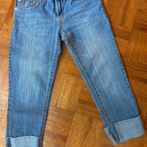 May include: Light blue denim capri jeans with a cuffed hem. The jeans have a classic five-pocket design, a button and zipper closure, and a small American flag detail on the front pocket. The jeans are displayed on a wooden floor.