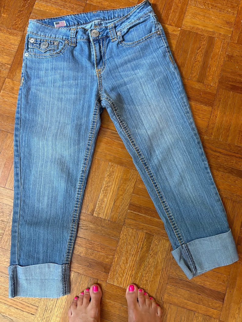 May include: Light blue denim capri jeans with a cuffed hem. The jeans have a classic five-pocket design, a button and zipper closure, and a small American flag detail on the front pocket. The jeans are displayed on a wooden floor.