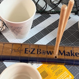 Op de afbeelding: Een houten Deluxe E-Z Bow Maker gereedschap met een bedrukte liniaal, blauwe accenten en drie verticale houten deuvels. Het apparaat bevat de tekst Deluxe E-Z Bow Maker en E-Z Craft. Het ligt op een rastermat naast witte keramische mokken.