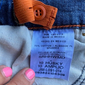 Op de afbeelding: Een close-up van een blauw denim jeanslabel met witte tekst die "RN 130273 MADE IN MEXICO HECHO EN MEXICO 99% COTTON / ALGODON 1% SPANDEX EXCLUSIVE OF DECORATION! EXCEPTO DECORACION 5RHHWMD 10 HUSKY 10 AMPLIO JIM-L091-014 083624104309" luidt.