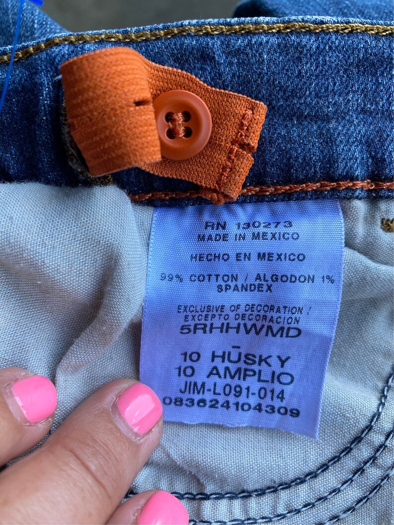 Op de afbeelding: Een close-up van een blauw denim jeanslabel met witte tekst die "RN 130273 MADE IN MEXICO HECHO EN MEXICO 99% COTTON / ALGODON 1% SPANDEX EXCLUSIVE OF DECORATION! EXCEPTO DECORACION 5RHHWMD 10 HUSKY 10 AMPLIO JIM-L091-014 083624104309" luidt.