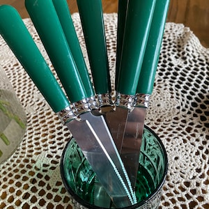 May include: A set of six green handled butter knives with silver blades are arranged in a green glass jar. The jar is sitting on a white crocheted doily.
