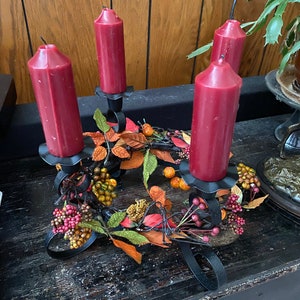 May include: A black metal candle holder with four red candles and fall-themed decorations. The holder has a circular base with a wreath of faux leaves and berries.