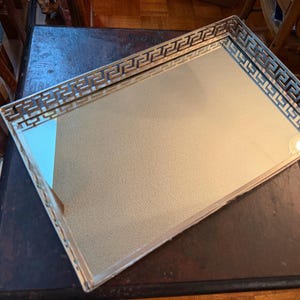 May include: A rectangular mirrored tray featuring a metallic silver frame with an intricate openwork Greek key pattern border. The central surface is a clear, reflective glass panel, providing a smooth and polished finish for this decorative item.