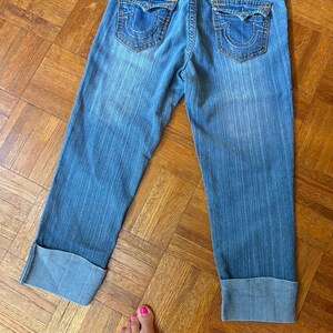 May include: Light blue denim capri jeans with a faded wash. Features include large back pockets with decorative stitching, a small logo patch, and folded cuffs. The jeans are laid flat on a wooden floor.