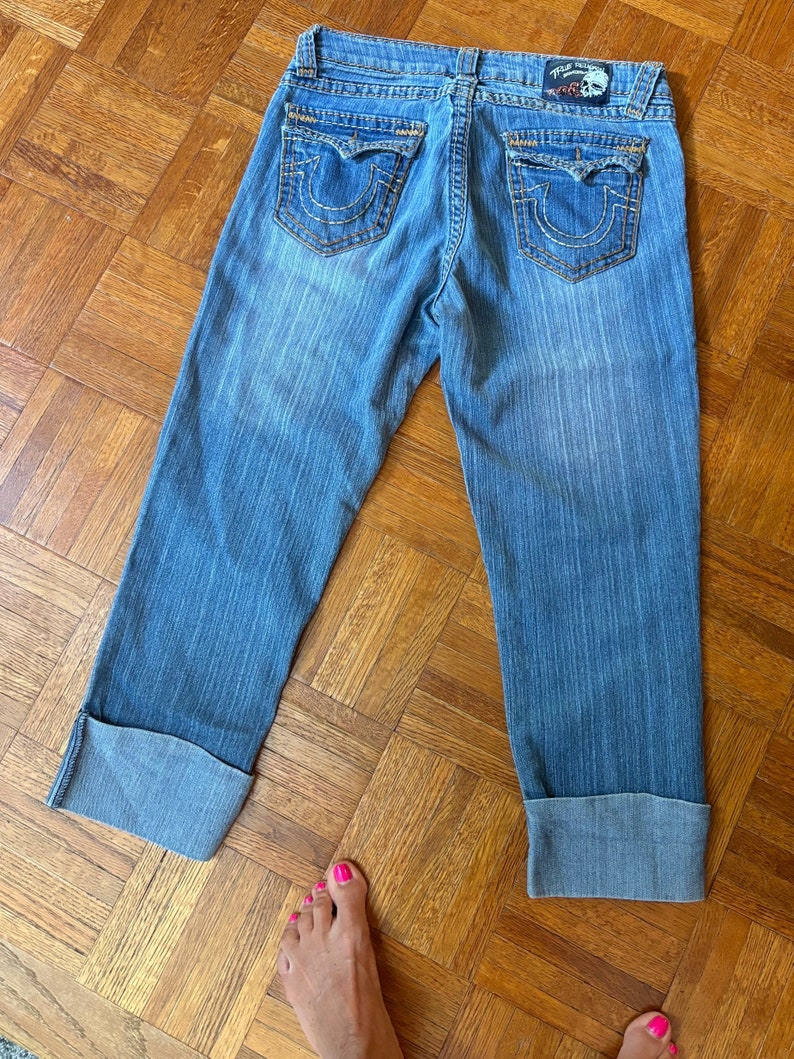 May include: Light blue denim capri jeans with a faded wash. Features include large back pockets with decorative stitching, a small logo patch, and folded cuffs. The jeans are laid flat on a wooden floor.