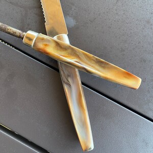 May include: Two knives with brown and white marbled handles. The knives are crossed over each other. The knife on top has a serrated blade.