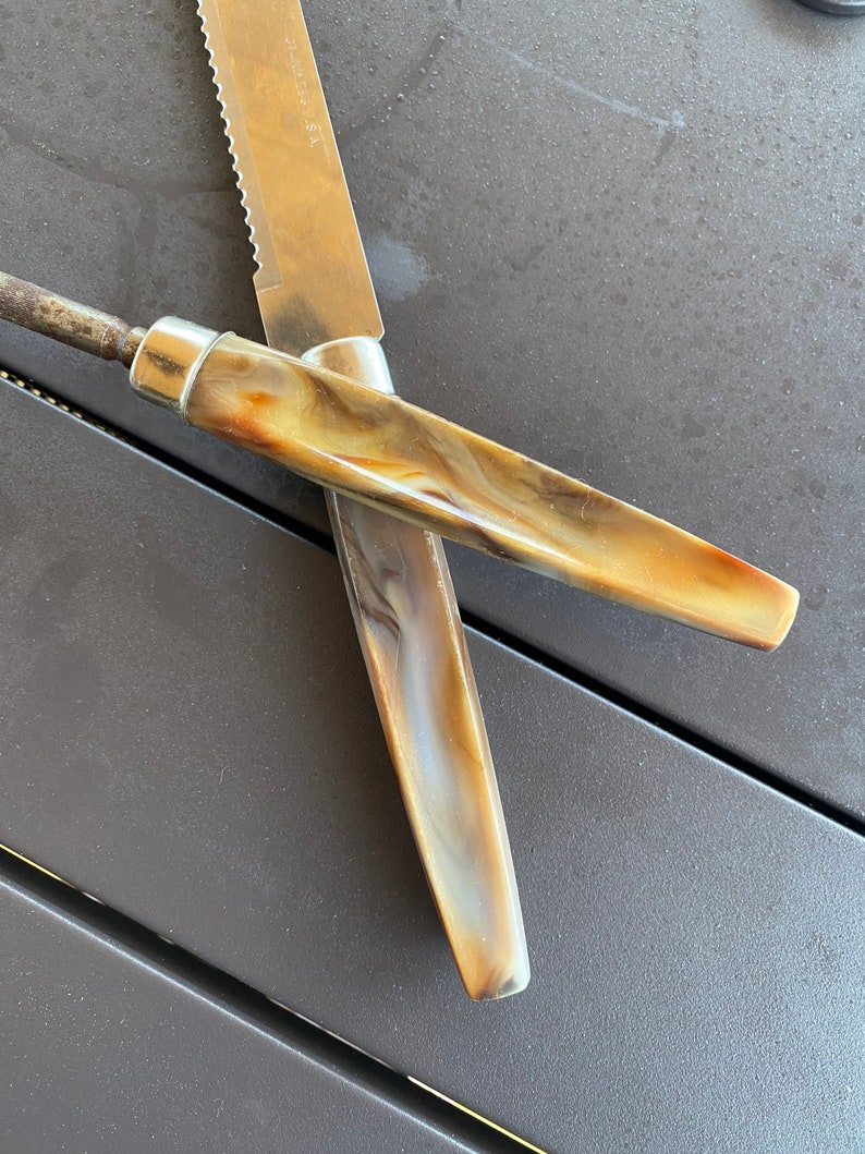 May include: Two knives with brown and white marbled handles. The knives are crossed over each other. The knife on top has a serrated blade.