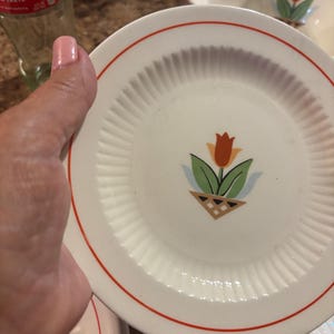 Puede incluir: Plato de cerámica blanco con borde rojo y diseño de tulipán en el centro. El tulipán es naranja y rojo con hojas verdes y está en una cesta marrón. El plato tiene una textura acanalada.