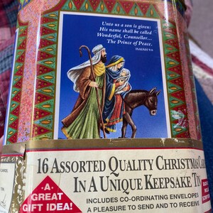 Puede incluir: Una lata rectangular para tarjetas navideñas con un marco dorado y una tapa decorativa. La lata presenta una escena bíblica con el texto "Unto us a son is given". La lata dice "16 Assorted Quality Christmas Cards in a Unique Keepsake Tin."