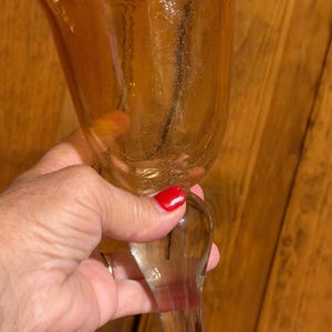 May include: A clear glass goblet with a wide, amber-colored bowl and a clear stem. The goblet has a flared rim and a rounded base. The glass has a slightly textured appearance. The goblet is held in a hand.