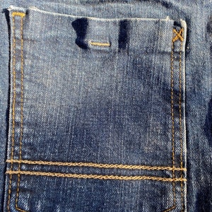 Op de afbeelding: Een close-up van een blauwe denim achterzak met een dubbele gestikte bruine draaddetail en een zwart "Wrangler" label.