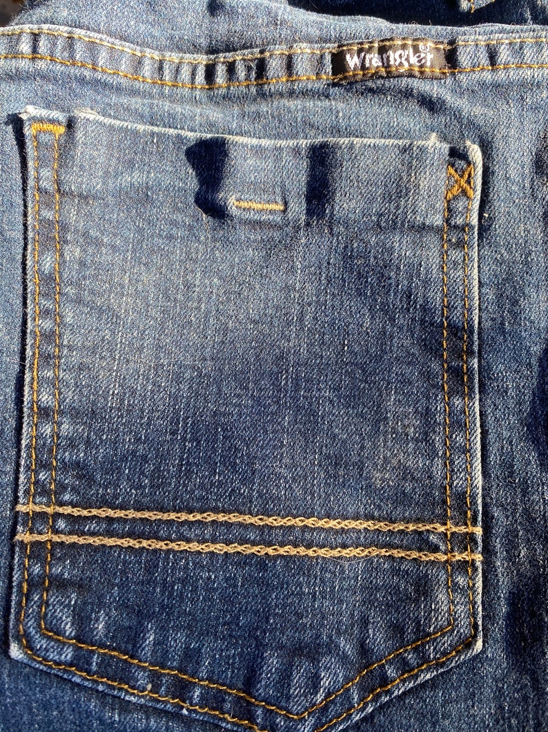 Op de afbeelding: Een close-up van een blauwe denim achterzak met een dubbele gestikte bruine draaddetail en een zwart "Wrangler" label.
