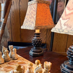 May include: A small table lamp with a pineapple-shaped base and an orange and beige patterned lampshade. The lamp sits on a dark wooden surface, with a chess set in the foreground. The lamp is plugged in and lit.