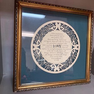 May include: A square wall art piece in a gold ornate frame featuring a teal background and a circular cream-colored paper cutout. The cutout displays the text: Love is patient, love is kind and is not jealous, love does not brag, and is not arrogant, does not seek its own, is not provoked, does not take into account a wrong suffered, LOVE does not rejoice in unrighteousness, but rejoices with the truth, bears all things, believes all things, hopes all things, endures all things. Love never fails. 1 Corinthians 13:4-8.