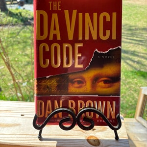 May include: A red paperback book with the title "The Da Vinci Code" in gold lettering. The book is a novel by Dan Brown, author of "Angels & Demons".