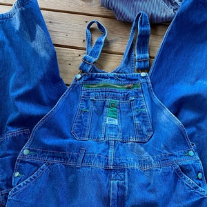 Könnte beinhalten: Blaue Jeans-Latzhosen mit grünem Besatz und einer Reißverschlusstasche. Die Latzhosen haben verstellbare Träger und mehrere Taschen.