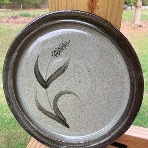 Könnte beinhalten: Ein runder Keramikteller mit einem dunkelbraunen Rand und einer hellgrauen, gesprenkelten Glasur. Ein schwarzes Blumenmuster ist auf den Teller gemalt.