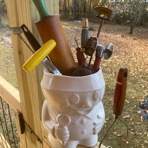 Puede incluir: Una figurita de cerámica blanca de una persona sosteniendo una cuchara, con una variedad de utensilios de cocina, incluyendo un rodillo, un machacador de patatas y un termómetro para carne, dentro de la figurita.