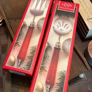 Könnte beinhalten: Zwei Lenox Servierutensilien in roten Schachteln. Eines ist eine Gabel, das andere ein Schlitzlöffel. Beide haben silberne Metallköpfe und rote Griffe. Die Schachteln sind rot mit dem Lenox-Logo und dem Schriftzug "American by Design".