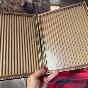May include: A vintage gold-toned double picture frame. The frame is rectangular with a decorative rope-like border. The interior features a tan, vertically-lined backing. The frame is open, revealing the glass and backing.