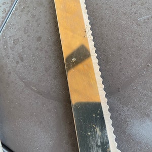 May include: A close-up of a serrated knife blade with a shiny, metallic finish. The blade has a slight curve and is partially covered in a reflection of a checkered pattern.