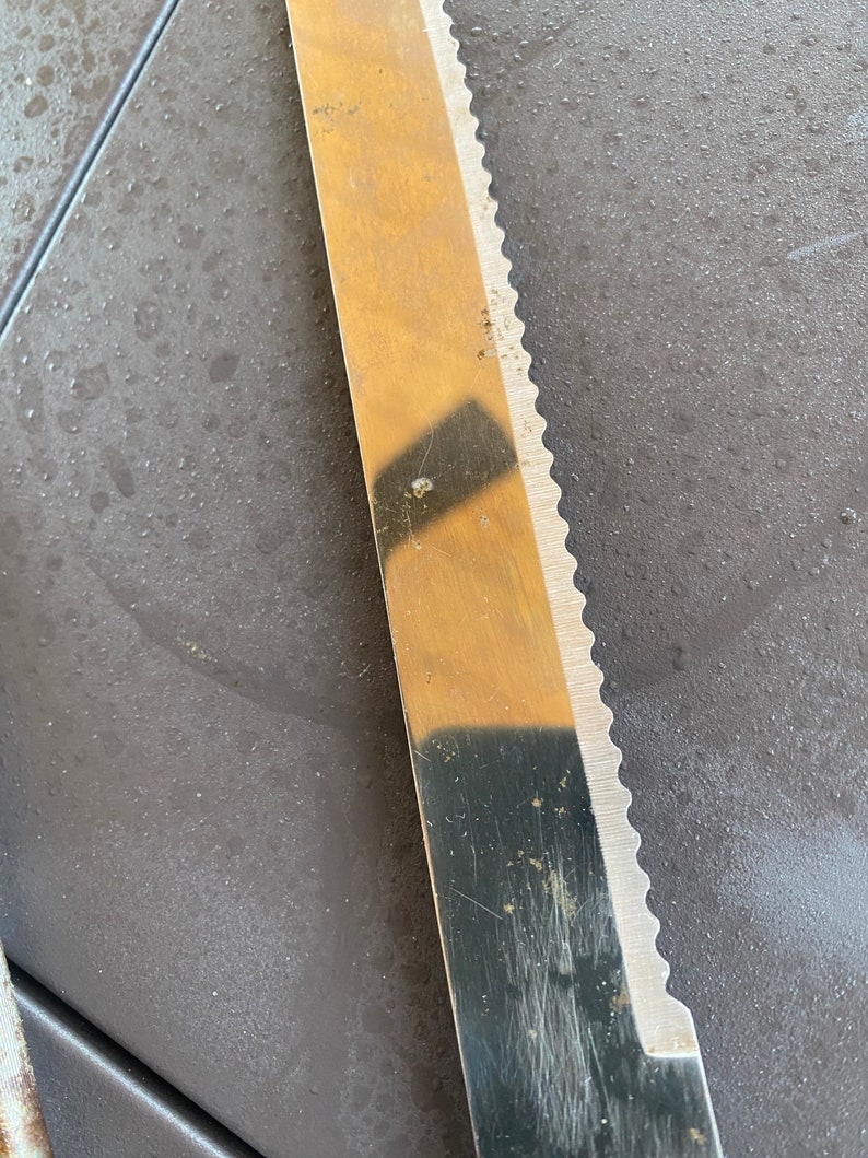 May include: A close-up of a serrated knife blade with a shiny, metallic finish. The blade has a slight curve and is partially covered in a reflection of a checkered pattern.