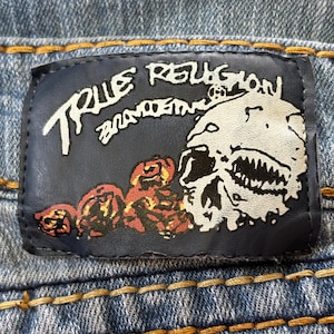 May include: Close-up of a blue denim jeans label. The label is dark blue with white text that reads "TRUE RELIGION BRAND JEANS". The label features a skull graphic and red and brown floral designs.