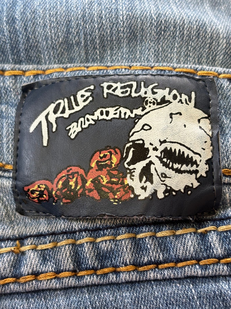 May include: Close-up of a blue denim jeans label. The label is dark blue with white text that reads "TRUE RELIGION BRAND JEANS". The label features a skull graphic and red and brown floral designs.