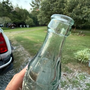 Puede incluir: Una botella de vidrio transparente vintage con una forma contorneada distintiva y un borde grueso y redondeado. El vidrio tiene un ligero tono verdoso y presenta letras en relieve y detalles texturizados a lo largo del cuerpo.