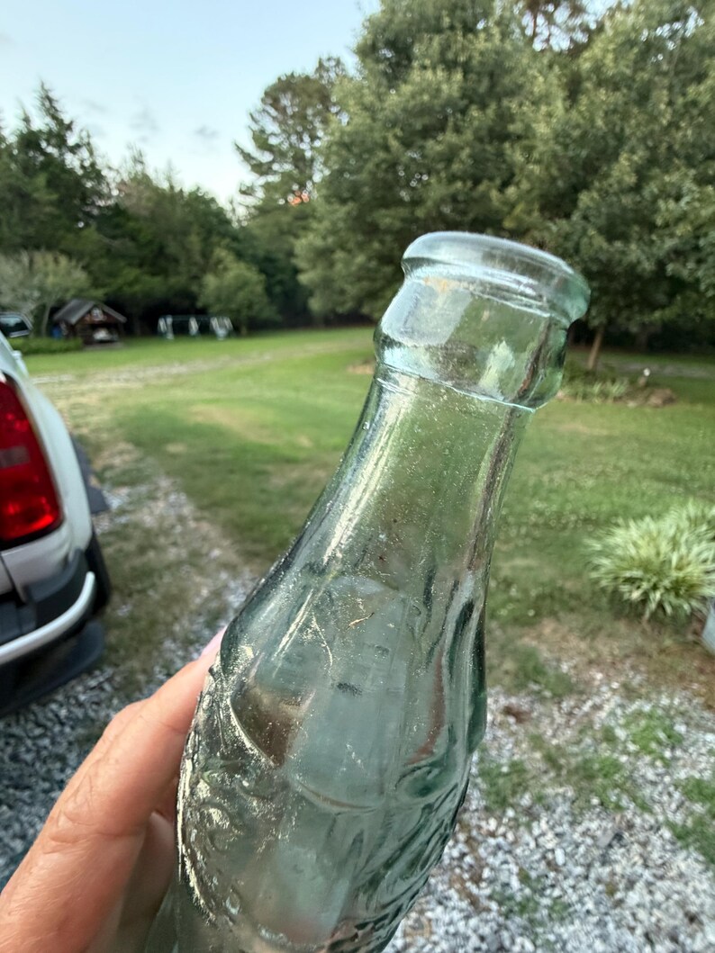 Puede incluir: Una botella de vidrio transparente vintage con una forma contorneada distintiva y un borde grueso y redondeado. El vidrio tiene un ligero tono verdoso y presenta letras en relieve y detalles texturizados a lo largo del cuerpo.