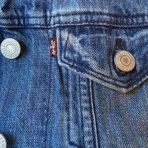 Op de afbeelding: Close-up van een blauw denim jack met twee zilveren knopen en een rood Levi's-label. Het jack heeft een zak aan de linkerkant.