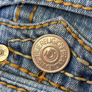 May include: Close-up of blue denim jeans with gold stitching. A silver button with the text "TRUE RELIGION BRAND JEANS" is visible. The jeans have a classic five-pocket design.