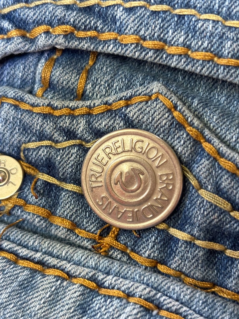 May include: Close-up of blue denim jeans with gold stitching. A silver button with the text "TRUE RELIGION BRAND JEANS" is visible. The jeans have a classic five-pocket design.